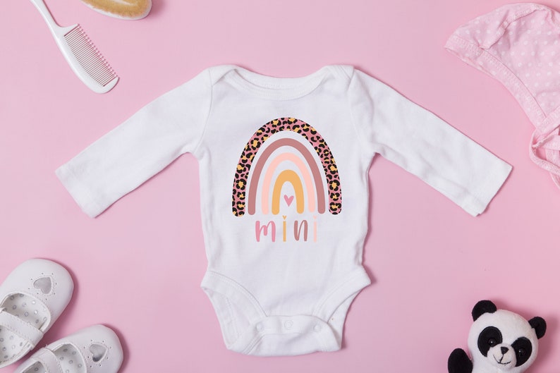 K&ouml;nnte beinhalten: Wei&szlig;er lang&auml;rmliger Baby-Strampler mit einem rosa, orangefarbenen und gelben Regenbogen mit Leopardenmuster. Der Regenbogen hat ein kleines pinkfarbenes Herz in der Mitte. Das Wort "mini" ist unter dem Regenbogen gedruckt.