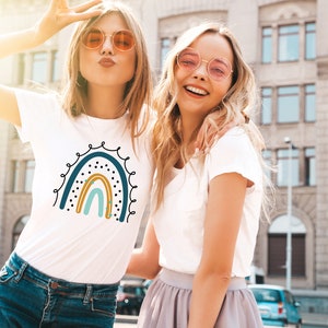 Op de afbeelding: Twee vriendinnen dragen witte T-shirts met een regenboog-print. De regenboog is blauw, turquoise, geel en oranje met zwarte stippen en gouden sterren. De vriendinnen lachen en dragen zonnebrillen.