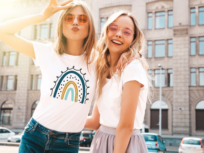 Op de afbeelding: Twee vriendinnen dragen witte T-shirts met een regenboog-print. De regenboog is blauw, turquoise, geel en oranje met zwarte stippen en gouden sterren. De vriendinnen lachen en dragen zonnebrillen.
