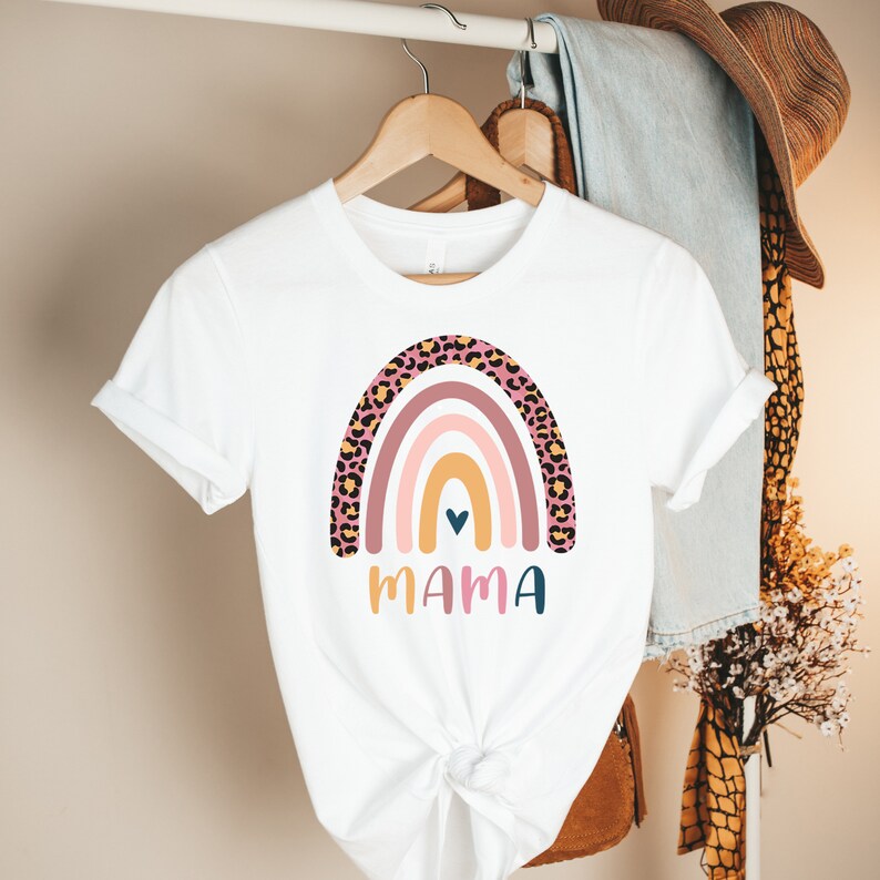 K&ouml;nnte beinhalten: Wei&szlig;es T-Shirt mit einem Regenbogen-Design mit rosa, gelben und orangen Streifen. Der Regenbogen ist mit einem Leopardenmuster umrandet. Das Wort "MAMA" ist unter dem Regenbogen gedruckt.