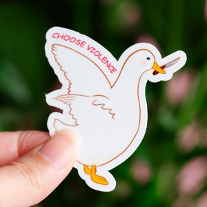 May include: A white sticker featuring a cartoon duck holding a knife in its beak. The sticker has a brown outline and yellow feet. The text "CHOOSE VIOLENCE" is printed in red above the duck.