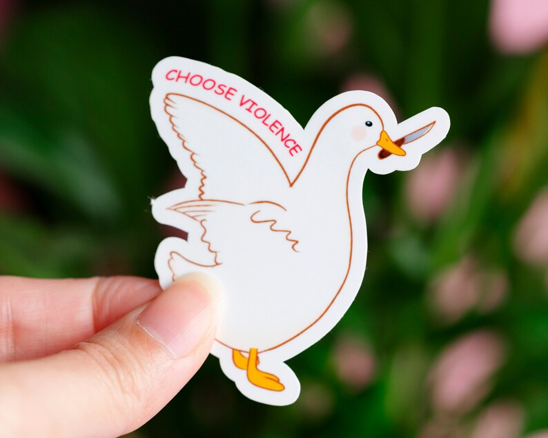 May include: A white sticker featuring a cartoon duck holding a knife in its beak. The sticker has a brown outline and yellow feet. The text "CHOOSE VIOLENCE" is printed in red above the duck.