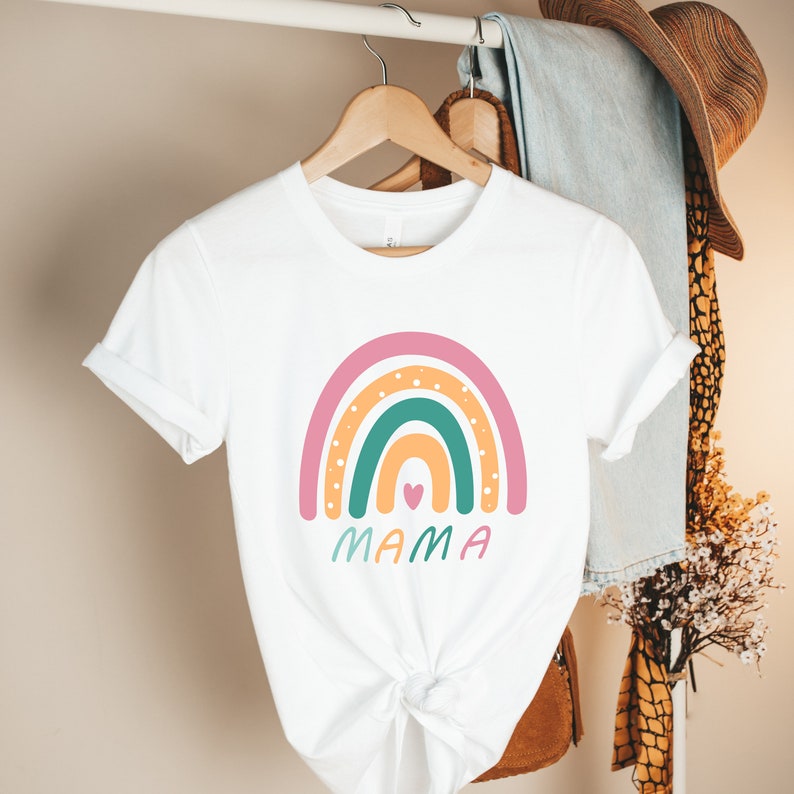 Op de afbeelding: Wit T-shirt met een regenboogontwerp en het woord "MAMA" eronder gedrukt. De regenboog is gemaakt van de kleuren roze, oranje, geel, groen en blauw met witte stippen.