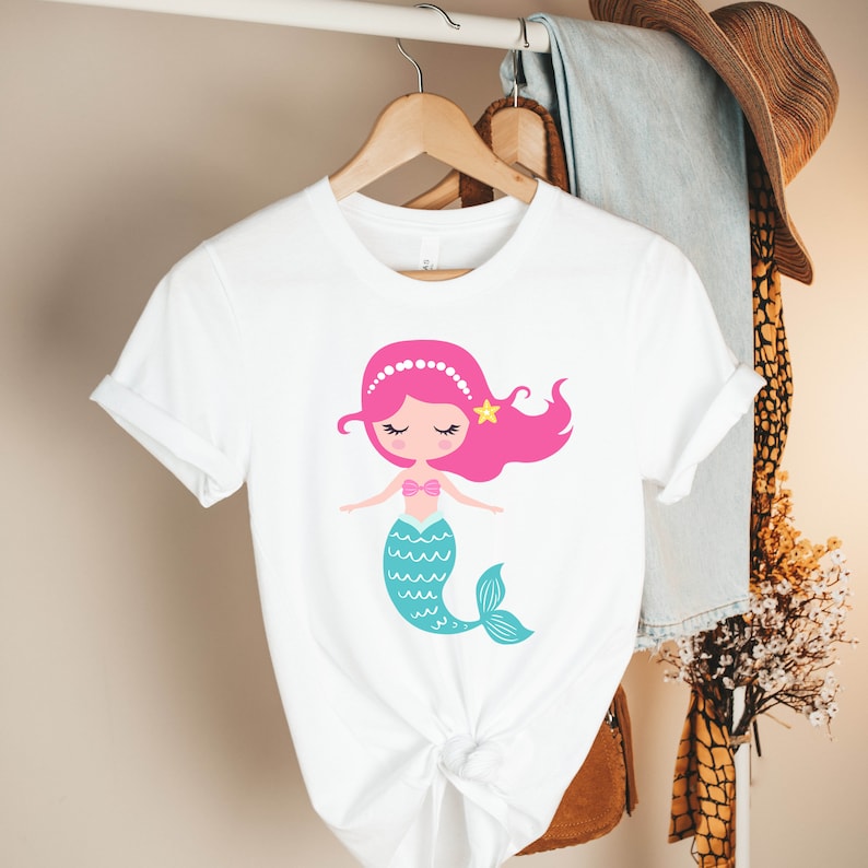 May include: White t-shirt with a cartoon mermaid design. The mermaid has pink hair, a blue tail, and a pink seashell bra. She is wearing a pearl headband and a yellow star.