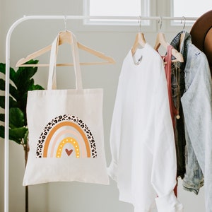 K&ouml;nnte beinhalten: Eine wei&szlig;e Canvas-Tragetasche mit einem Regenbogen-Design mit braunen und schwarzen Leopardenmuster-Akzenten. Der Regenbogen hat ein Herz in der Mitte und ist in Orange-, Gelb-, Rosa- und Wei&szlig;t&ouml;nen gehalten.