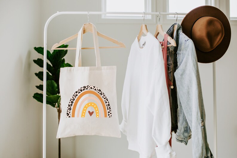 K&ouml;nnte beinhalten: Eine wei&szlig;e Canvas-Tragetasche mit einem Regenbogen-Design mit braunen und schwarzen Leopardenmuster-Akzenten. Der Regenbogen hat ein Herz in der Mitte und ist in Orange-, Gelb-, Rosa- und Wei&szlig;t&ouml;nen gehalten.