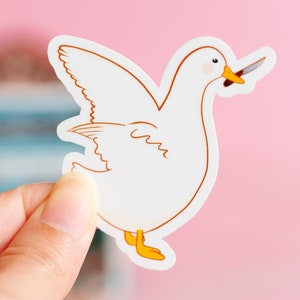 May include: A white sticker featuring a cartoon goose with a knife in its beak. The goose has brown outlines, yellow feet, and a pink blush on its cheeks. The sticker is held by a hand against a pink background.