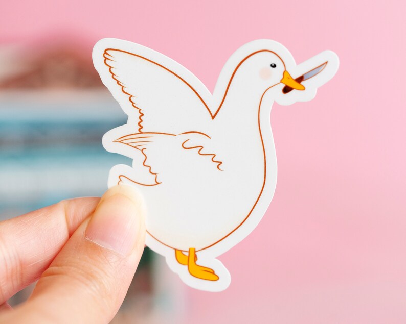 May include: A white sticker featuring a cartoon goose with a knife in its beak. The goose has brown outlines, yellow feet, and a pink blush on its cheeks. The sticker is held by a hand against a pink background.