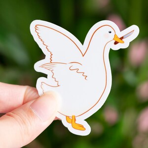 May include: A white sticker featuring a cartoon goose with a knife in its beak. The goose has orange feet and a yellow beak. The sticker is held against a blurred green and pink background.