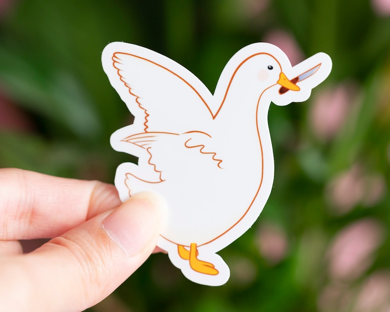 May include: A white sticker featuring a cartoon goose with a knife in its beak. The goose has orange feet and a yellow beak. The sticker is held against a blurred green and pink background.