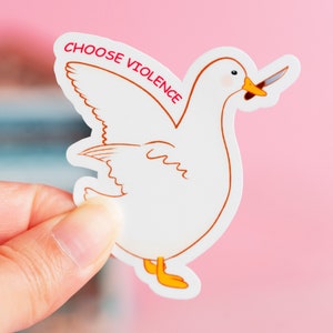 May include: A white sticker featuring a cartoon duck with a knife in its beak. The duck has orange feet and a brown outline. The text "CHOOSE VIOLENCE" is printed in red above the duck.