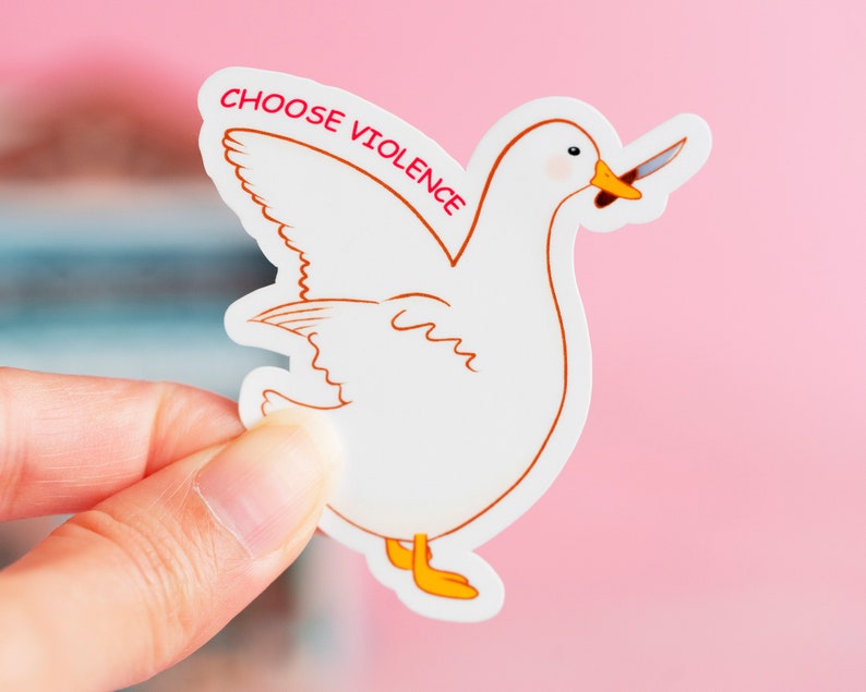 May include: A white sticker featuring a cartoon duck with a knife in its beak. The duck has orange feet and a brown outline. The text "CHOOSE VIOLENCE" is printed in red above the duck.
