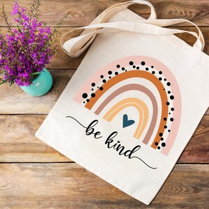 K&ouml;nnte beinhalten: Eine wei&szlig;e Canvas-Tragetasche mit einem Regenbogen-Design und dem Text "be kind". Der Regenbogen besteht aus orangefarbenen, braunen, rosa und gelben Streifen mit schwarzen Punkten. Ein blaues Herz befindet sich in der Mitte des Regenbogens.