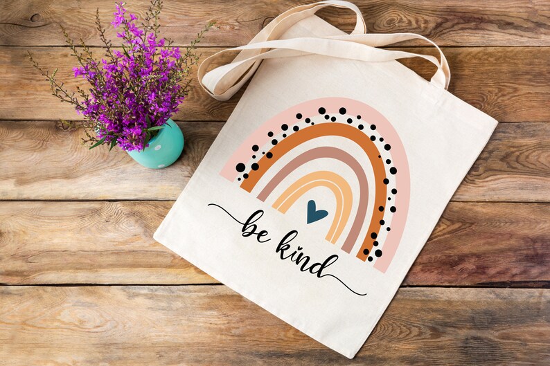 K&ouml;nnte beinhalten: Eine wei&szlig;e Canvas-Tragetasche mit einem Regenbogen-Design und dem Text "be kind". Der Regenbogen besteht aus orangefarbenen, braunen, rosa und gelben Streifen mit schwarzen Punkten. Ein blaues Herz befindet sich in der Mitte des Regenbogens.