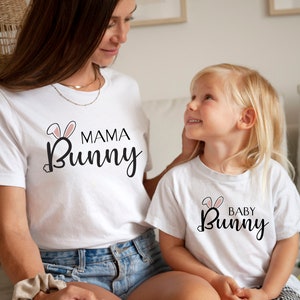 Può includere: Una maglietta bianca con il testo "Mama Bunny" in scrittura nera con orecchie di coniglio rosa. Una seconda maglietta bianca con il testo "Baby Bunny" in scrittura nera con orecchie di coniglio rosa. Le magliette sono indossate da una donna e un bambino.