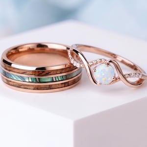 May include: A rose gold band featuring a central inlay of wood and iridescent abalone shell, paired with a rose gold twisted band ring set with a round white opal stone and small clear accent stones along the band's delicate, intertwined design.