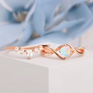 May include: Two rose gold rings. One ring has a curved design with clear gemstones. The other ring features an infinity-style band with a central opal-like stone and small accent stones. The rings are set against a soft, blurred background.