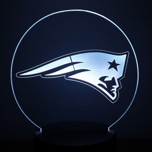 May include: A white LED night light shaped like a circle with the New England Patriots logo in the center. The logo is a white silhouette of a flying football helmet with a star on the side.