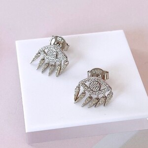 Può includere: Un paio di orecchini a lobo argentati. Ogni orecchino presenta un design a occhio con un cerchio centrale di piccole pietre trasparenti e una frangia di accenti appuntiti. Gli orecchini sono esposti su una superficie bianca su uno sfondo rosa tenue.