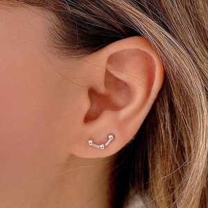 Può includere: Primo piano di un orecchio con un piccolo orecchino d'argento con una linea curva di tre perline collegate.