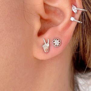 May include: A close-up of a person's ear with three silver earrings. The earrings are a peace sign, a flower, and a curved hoop with a small diamond.