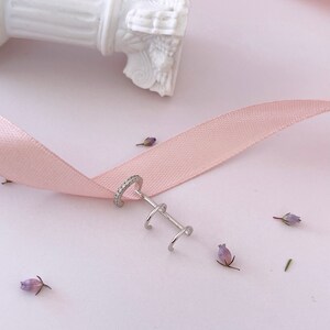 May include: Silver ear cuff with a single row of cubic zirconia stones and two smaller hoops. The ear cuff is on a pink ribbon and a white background.