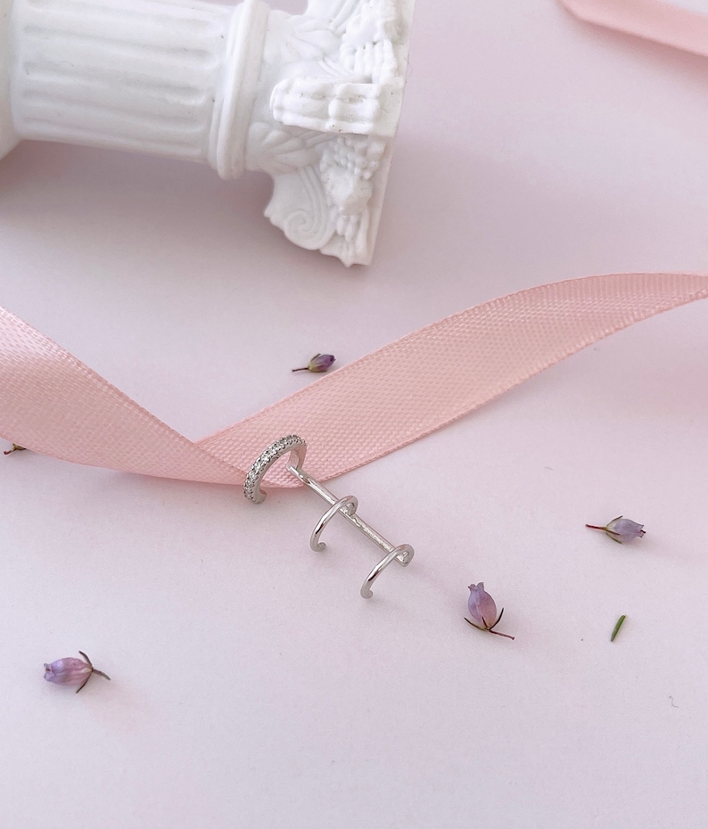 May include: Silver ear cuff with a single row of cubic zirconia stones and two smaller hoops. The ear cuff is on a pink ribbon and a white background.