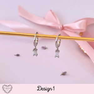 May include: Silver hoop earrings with a dangling rectangular clear stone. The earrings are displayed on a gold stand against a pink background. Design 1.