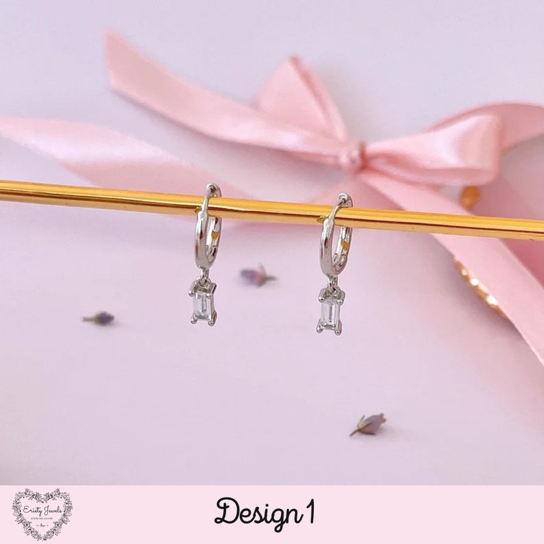 May include: Silver hoop earrings with a dangling rectangular clear stone. The earrings are displayed on a gold stand against a pink background. Design 1.