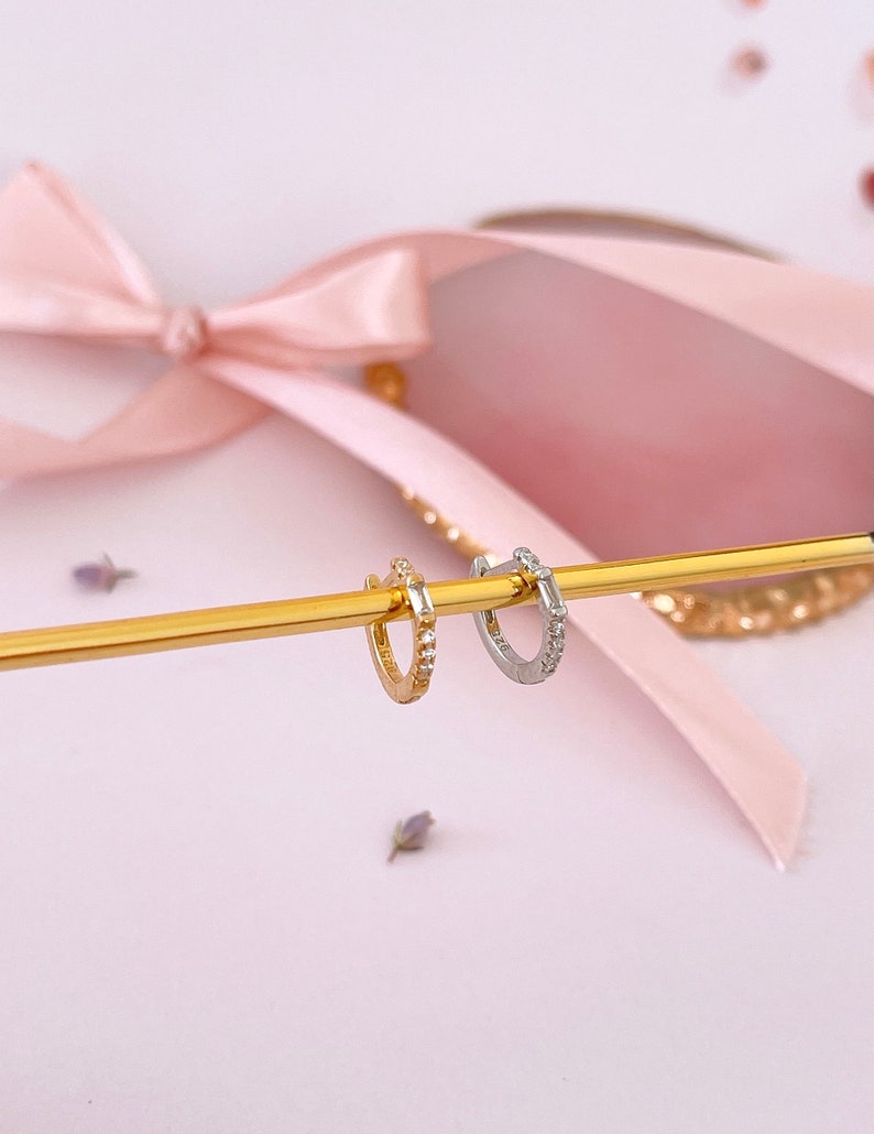 May include: Two small hoop earrings with a row of clear cubic zirconia stones. The hoop on the left is gold and the hoop on the right is silver.