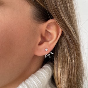 Puede incluir: Un par de pendientes de plata con un pequeño pendiente de diamante y una orejera geométrica. La orejera tiene forma de estrella con un borde puntiagudo.