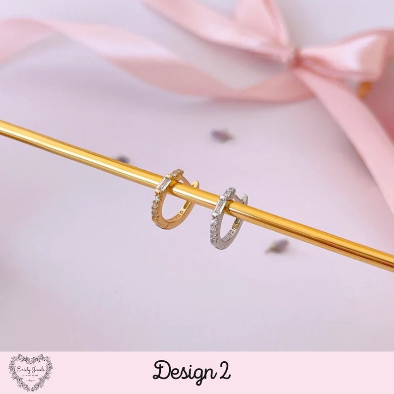 May include: Two small hoop earrings with a single row of small cubic zirconia stones and a larger, rectangular cubic zirconia stone on each hoop. The earrings are shown on a gold-colored surface. Design 2.