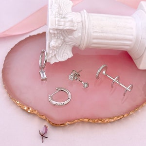 May include: A set of silver earrings on a pink and gold agate coaster. The earrings include a pair of small stud earrings with a single stone, a pair of hoop earrings with small stones, and a pair of hoop earrings with a single rectangular stone.