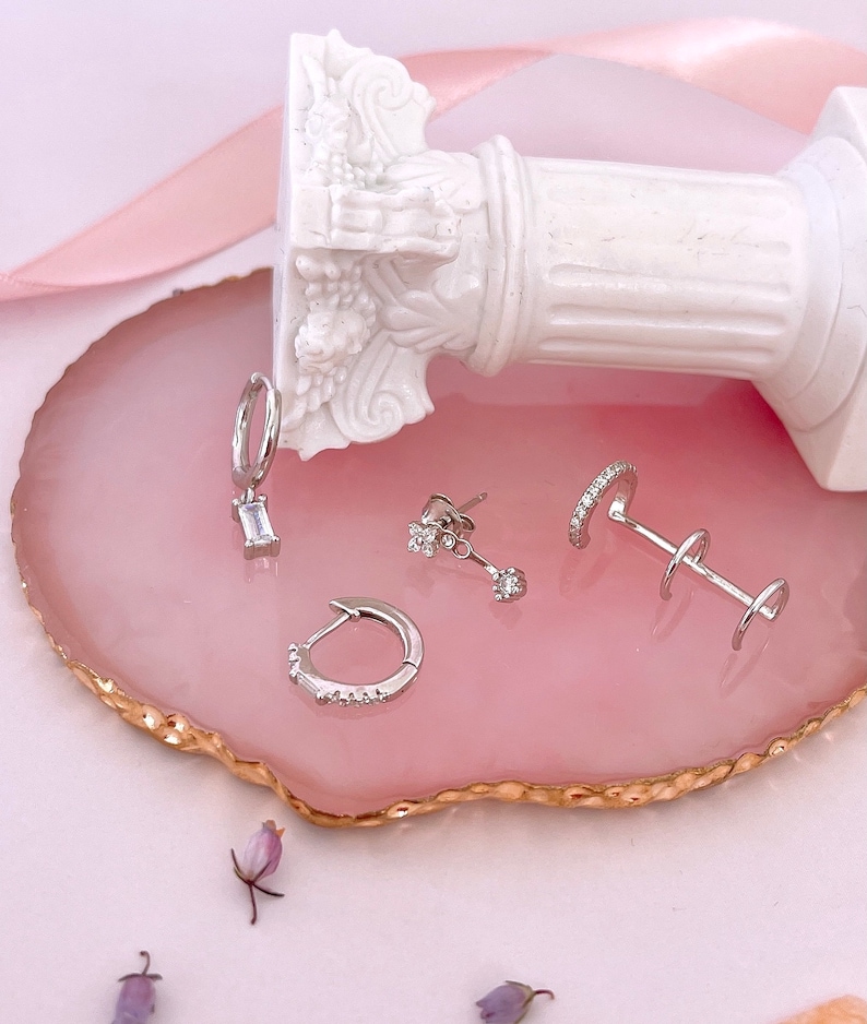 May include: A set of silver earrings on a pink and gold agate coaster. The earrings include a pair of small stud earrings with a single stone, a pair of hoop earrings with small stones, and a pair of hoop earrings with a single rectangular stone.