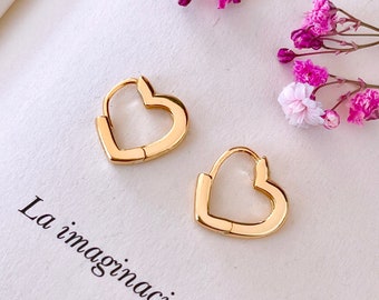 Pode incluir: Um par de brincos de argola de coração de ouro. Os brincos estão sobre uma superfície branca com um livro e flores cor-de-rosa no fundo.