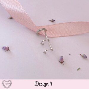 May include: Silver ear cuff with a triple hoop design and a single hoop with a row of small clear stones. The ear cuff is on a pink ribbon.
