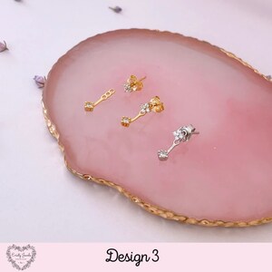 May include: Three pairs of earrings on a pink and gold agate-like surface. The earrings are gold and silver with small clear stones. The text "Design 3" is at the bottom of the image.