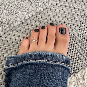 Peut inclure: Une bague d'orteil en argent avec un motif double chevron. La bague est portée sur le gros orteil d'un pied avec un vernis à ongles noir.