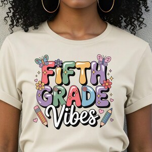 May include: A beige t-shirt with the words "FIFTH GRADE VIBES" in colourful bubble letters, decorated with butterflies, flowers, and pencils. The person is wearing blue jeans and a colourful headscarf.