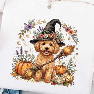 May include: A white crewneck sweatshirt featuring a graphic of a golden-colored dog wearing a black witch hat, holding a broomstick, and surrounded by orange pumpkins, colorful flowers, and small golden stars on a white background.