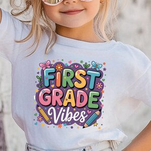 May include: A child wearing a white t-shirt with the text "FIRST GRADE VIBES" in colourful letters and graphics. The child is also wearing sunglasses and a large bow in their hair.