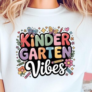 May include: White t-shirt featuring the text "Kindergarten Vibes" in a colourful design with butterflies, flowers, and pencils. The shirt is worn with denim shorts.