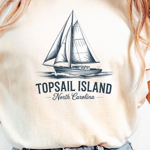 Puede incluir: Camiseta color crema con una ilustración detallada de un velero sobre el agua. El texto "TOPSAIL ISLAND - North Carolina" está impreso debajo en azul oscuro. La camiseta se combina con unos vaqueros azules.