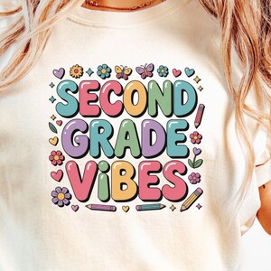 May include: A cream-coloured t-shirt featuring the text "SECOND GRADE VIBES" in a vibrant, retro style. The design incorporates flowers, butterflies, hearts, and pencils. The shirt is worn with light-wash blue jeans.