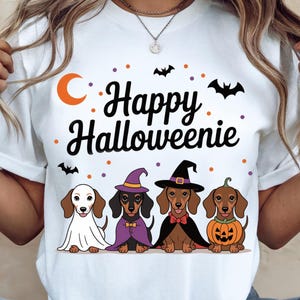 May include: White t-shirt with the text "Happy Halloweenie" and illustrations of four dachshunds in Halloween costumes. The dogs are dressed as a ghost, witch, vampire, and pumpkin. Orange moon and black bats complete the design.