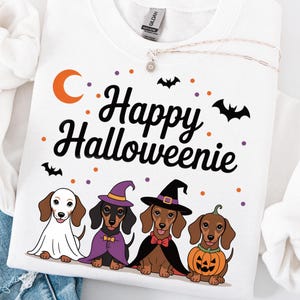 May include: White sweatshirt with the text "Happy Halloweenie" and illustrations of dachshunds in Halloween costumes. The dogs are dressed as a ghost, witch, vampire, and pumpkin. Orange moon and bat graphics.