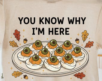 May include: Beige t-shirt with the text "YOU KNOW WHY I'M HERE" above a plate of deviled eggs. The graphic includes fall leaves and acorns. The shirt is hanging on a wooden hanger.