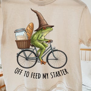 Puede incluir: Camiseta color crema con una ilustración caprichosa de una rana verde montando en bicicleta. La rana lleva un sombrero de mago marrón y lleva pan y un tarro en una cesta. El texto "OFF TO FEED MY STARTER" está impreso debajo.