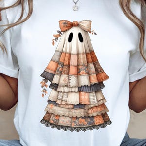 Può includere: T-shirt bianca con un design a fantasma stravagante. Il fantasma è adornato con un motivo patchwork nei toni dell'arancione, marrone e grigio, con un fiocco decorativo e accenti floreali. Una maglietta grafica unica a tema Halloween.