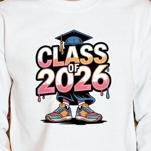 May include: White long-sleeve shirt with a colorful graphic that reads "Class of 2026." The design includes a graduation cap and stylized sneakers. The shirt is a casual style, suitable for graduation celebrations.